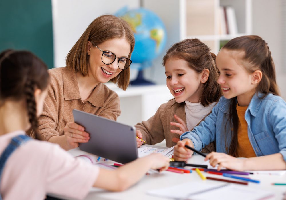Happy female teacher in glasses smiling and looking at little student while sitting at table and showing data on tablet during lesson at school